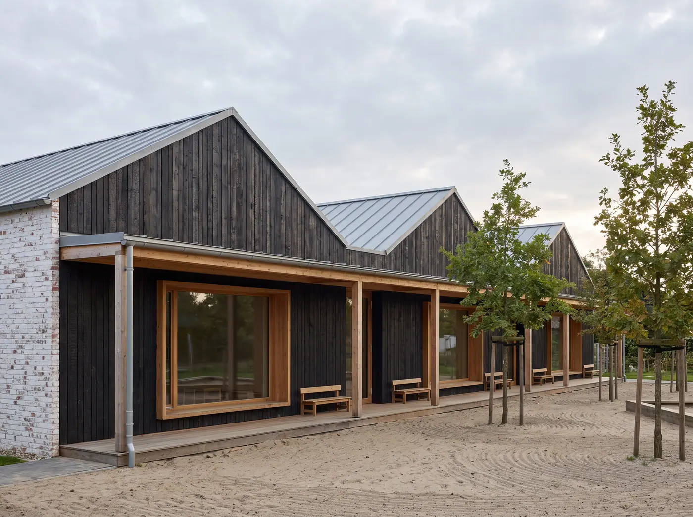 Kindertagesstätte aus drei Pavillons mit dunkler Holzfassade und Holzveranda am Sandspielplatz