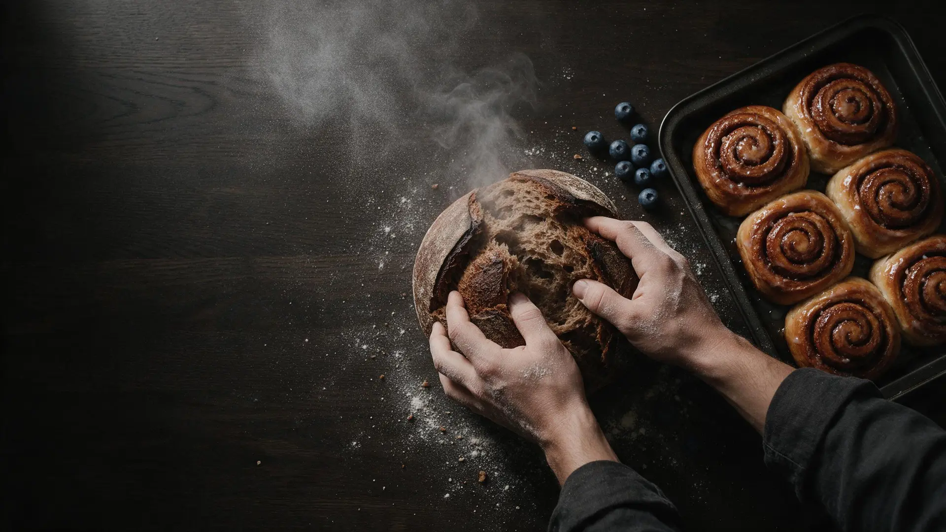Bemehlte Hände reißen ein dampfendes Roggenbrot auf, daneben ein Blech Zimtschnecken