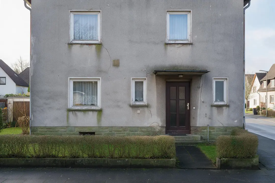 Graue, verwitterte Hausfassade mit grünen Algenspuren unter den Fensterbänken und Rissen im Putz
