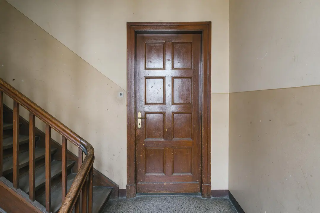 Altes Treppenhaus mit zerkratzter dunkelbrauner Wohnungstür und beigen, abgestoßenen Wänden