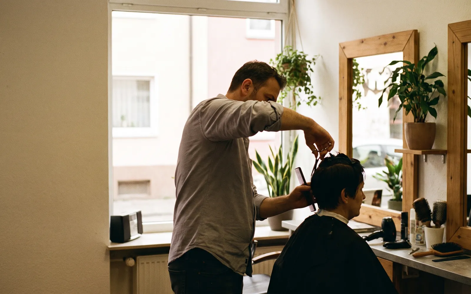 Friseur schneidet einer Kundin die Haare in einem kleinen Salon mit Holzspiegeln und Pflanzen