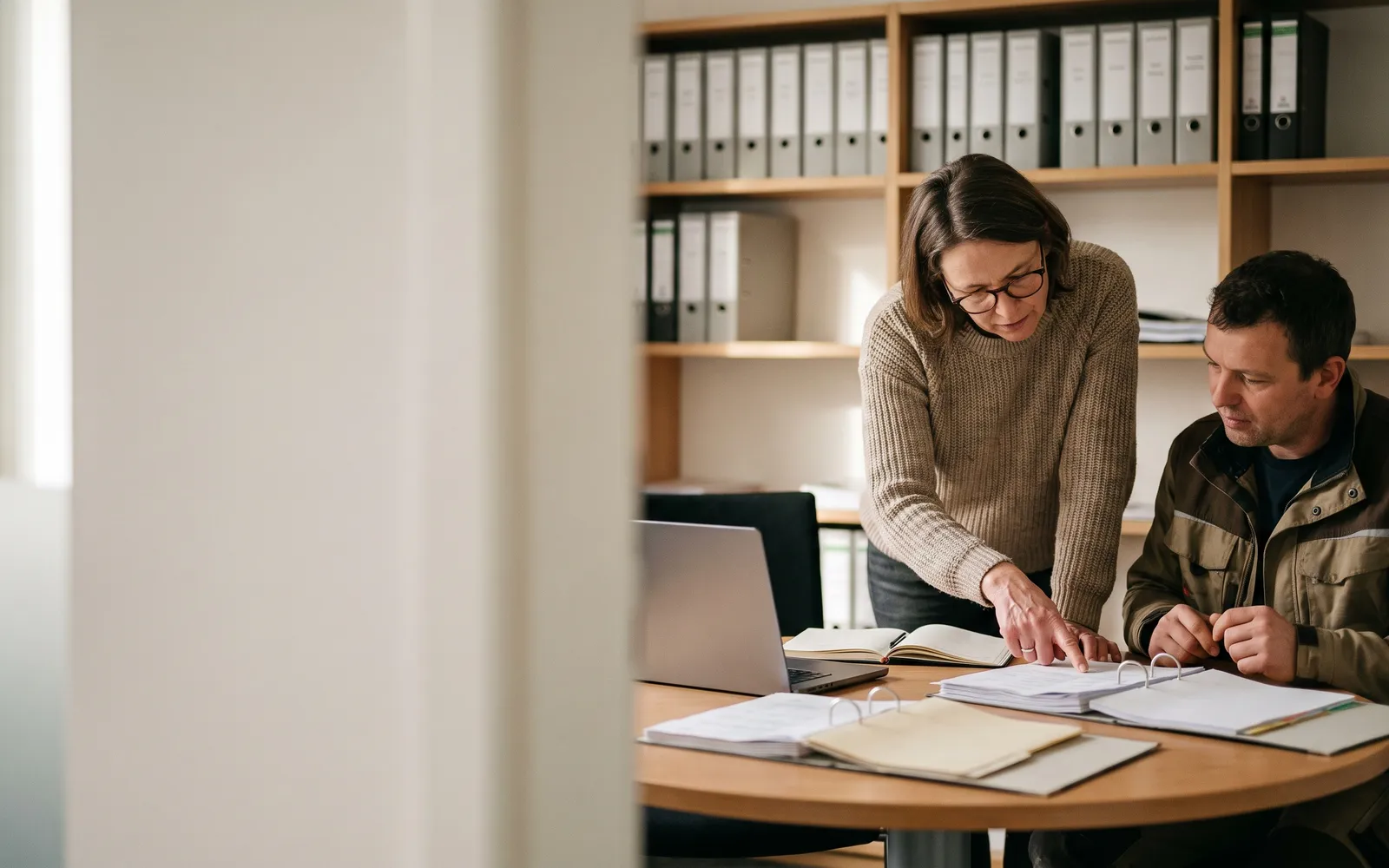 Steuerberaterin geht mit einem Handwerksunternehmer an einem runden Tisch Unterlagen und ein Notebook durch, im Hintergrund Regale mit Ordnern