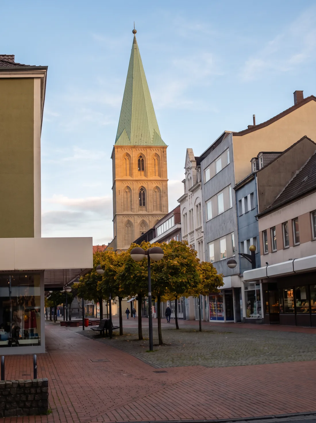 Die Pauluskirche in der Hammer Innenstadt im warmen Abendlicht: gotischer Sandsteinturm mit grüner Kupferspitze über der Fußgängerzone mit Bäumen.